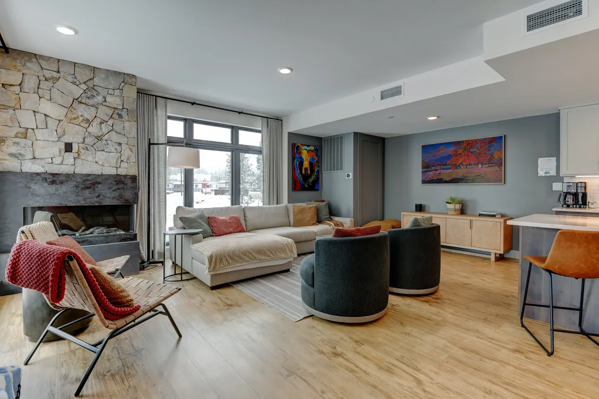 The image showcases a warm, inviting living room with a striking stone fireplace as the centerpiece, flanked by expansive windows revealing scenic mountain views. Modern art pieces adorn the walls, and plush seating is arranged for gathering, creating a space perfect for relaxation and conversation.