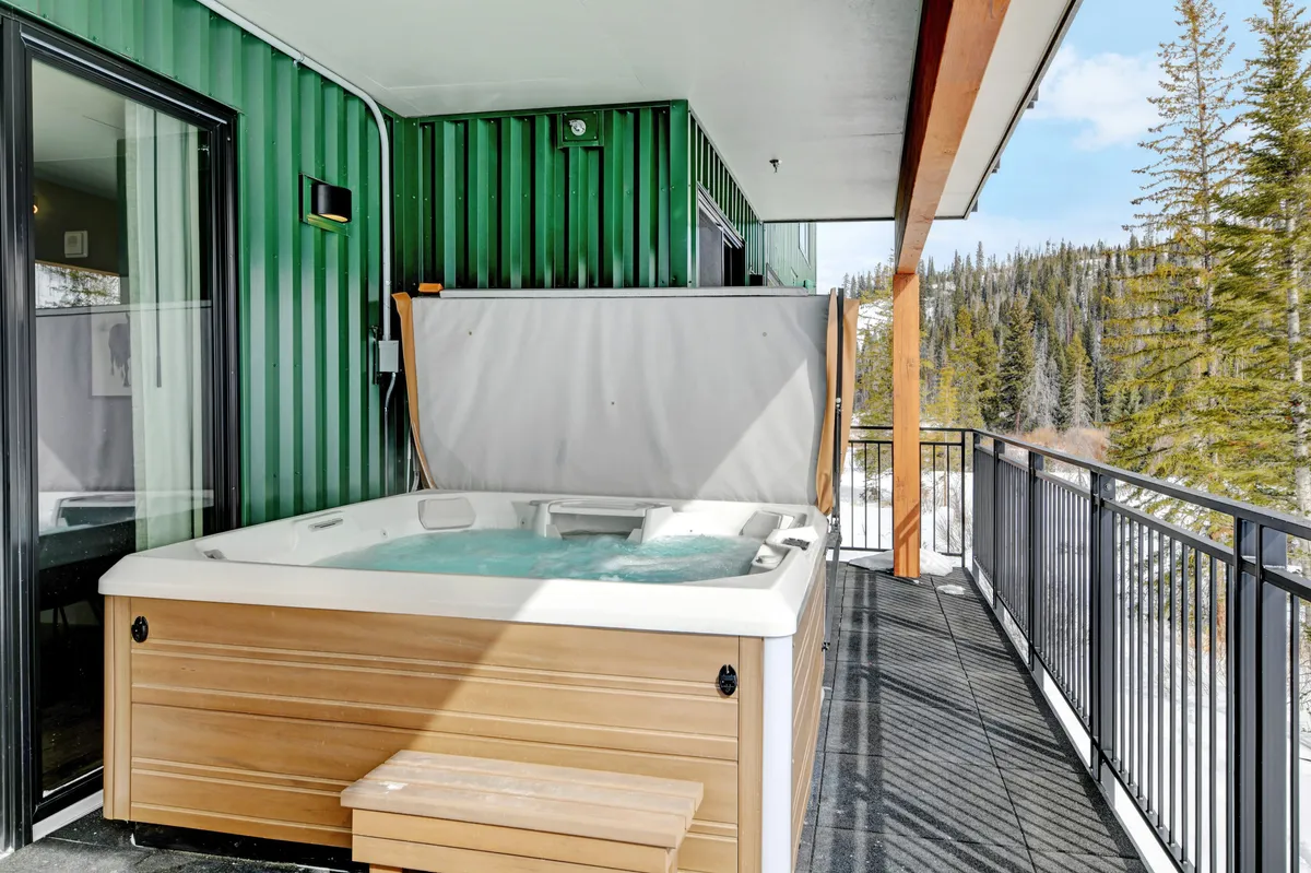 The image shows a modern hot tub set against a green wall on a wooden deck, with a view through the deck railing hinting at the expansive outdoors beyond. The scene feels tranquil and is well-suited for soaking at sunrise or sunset, surrounded by nature.