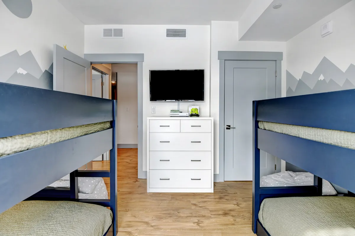 A playful bunk room featuring twin-over-twin bunk beds, a mountain mural, a white dresser, and a wall-mounted TV. The space is bright and whimsical, designed for fun sleepovers or movie marathons, complete with elements that appeal to both kids and the young-at-heart.