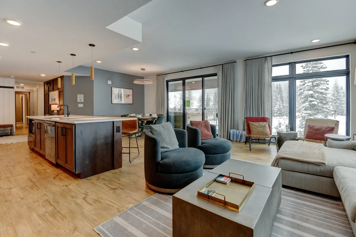 A bright, contemporary interior space with large windows showcasing snow-covered trees, a stylish kitchen island, and comfortable seating. The view outside hints at inspiring mountain vistas, while the interior suggests a cozy, work-friendly environment.