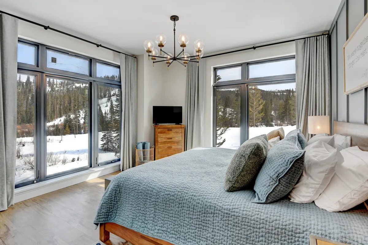 A spacious bedroom with a large bed featuring textured blue bedding, a stylish chandelier overhead, large windows framing a snowy landscape, and a wooden dresser. The scene radiates warmth and tranquility, showcasing a cozy spot to unwind and enjoy peaceful mornings or restful nights.