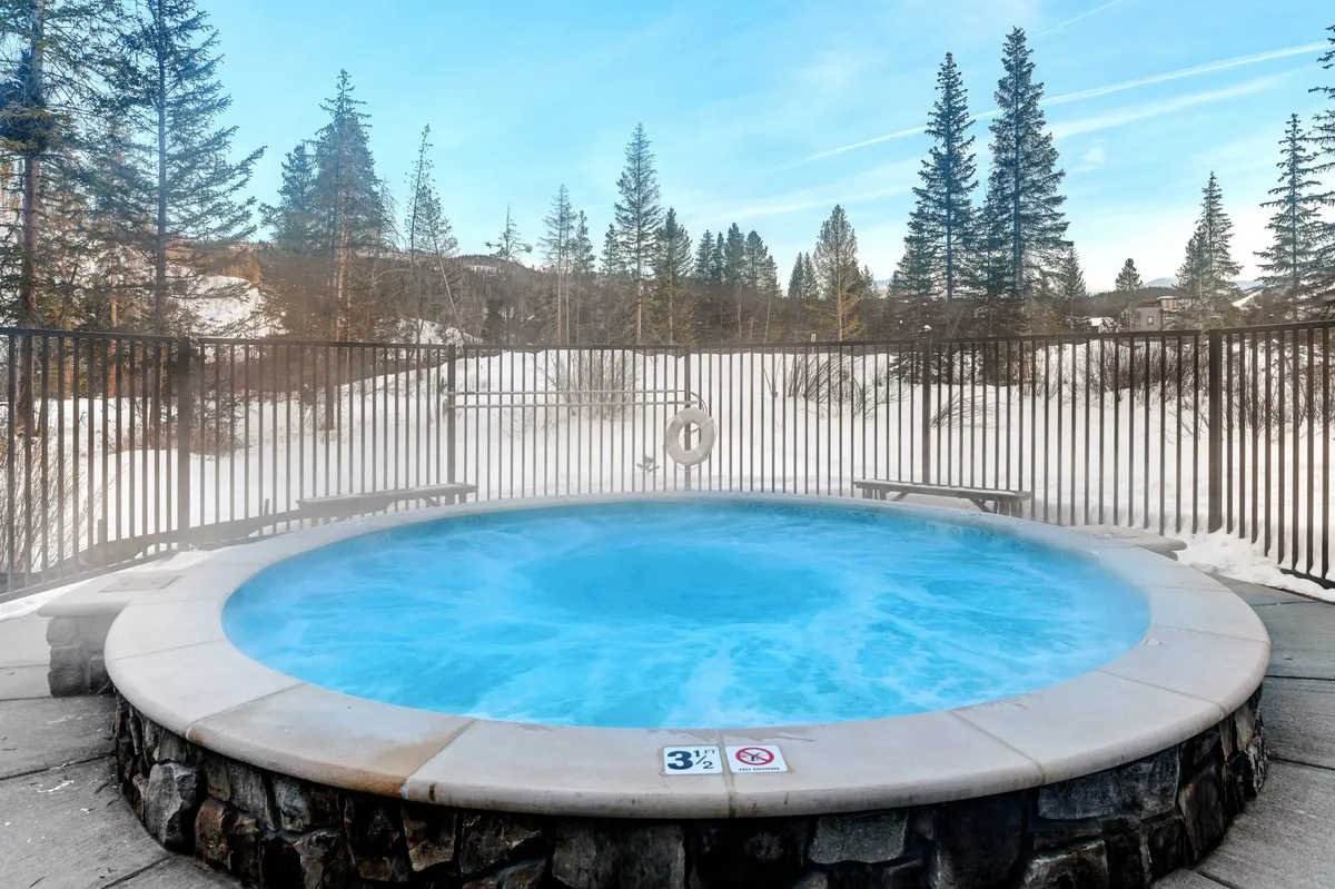 This exterior image features a hot tub set amidst a snowy landscape, surrounded by pine trees and enclosed by a fence. The contrast between the warm water and the crisp winter air is visually evident, inviting relaxation and enjoyment of the mountain environment.