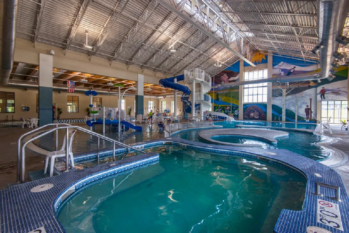 A vibrant indoor recreation center with a large swimming pool, colorful water slides, and children playing under a bright mural. The scene is energetic and family-friendly, capturing the excitement of offsite amenities like the Grand Park Rec Center.