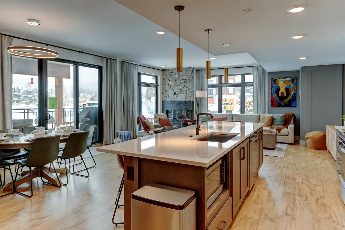 This image showcases an open-concept interior featuring a kitchen island, adjoining dining area, a cozy fireplace, large windows letting in natural light, and eye-catching artwork. The layout encourages interaction among guests, with spaces clearly designed for both cooking and relaxing.