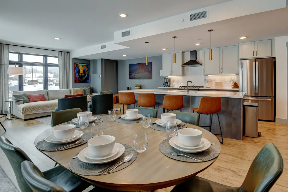 A warmly lit dining table is set for a meal, adjacent to a modern kitchen with stainless steel appliances and granite countertops. Large windows flood the space with natural light, creating an inviting atmosphere for gatherings.