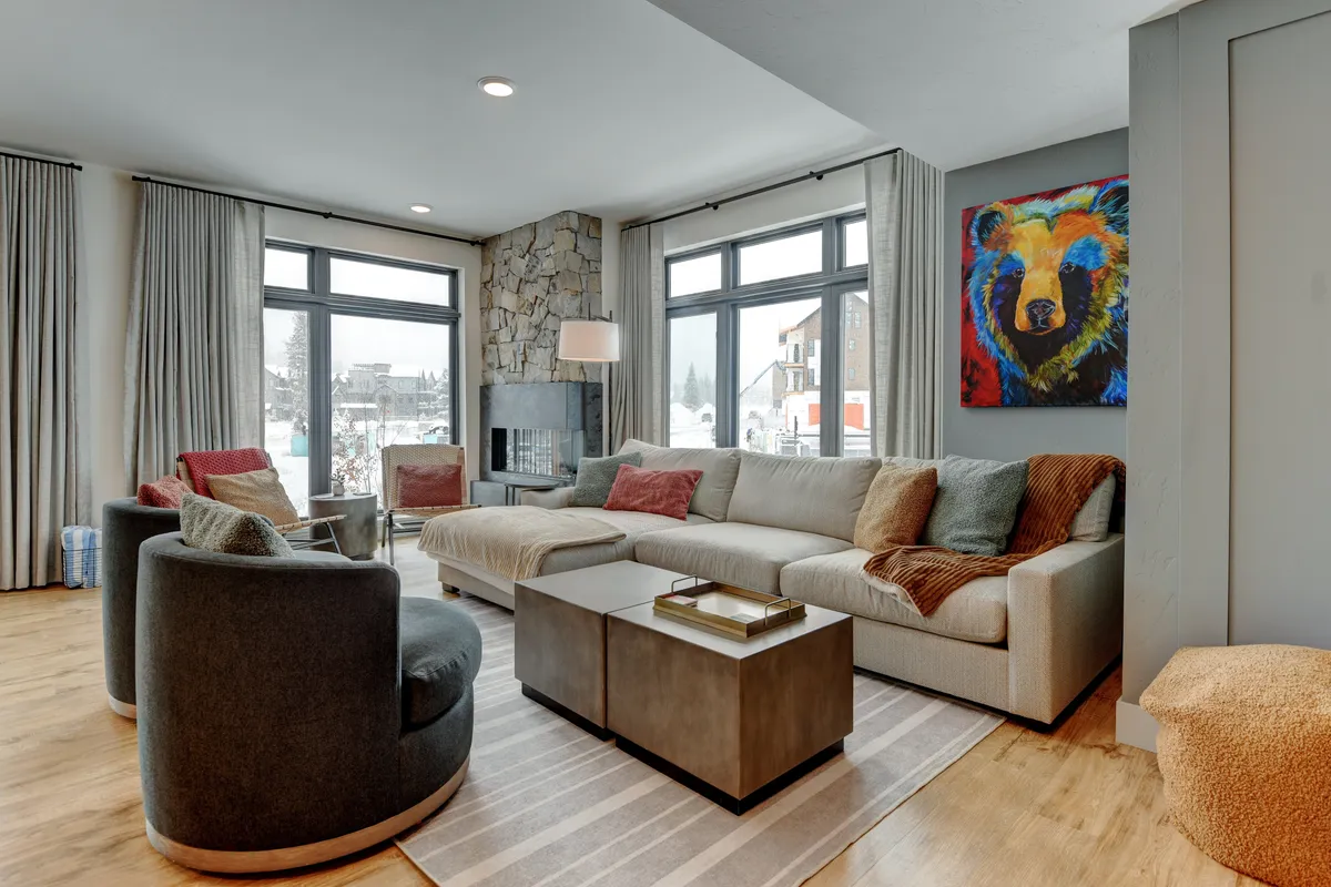 The living area features a large sectional sofa, a vibrant bear painting, a stone fireplace, and expansive windows with views. The setup is perfect for relaxing, socializing, or watching the 65” Frame TV, with plenty of space for board games or movie nights.