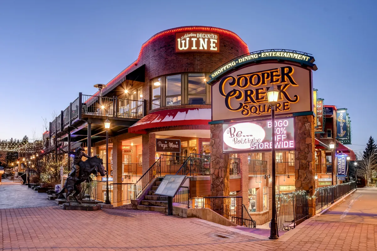 The image captures the Cooper Creek Square sign with prominent wine shop signage, inviting outdoor seating, and decorative lighting. The ambiance is lively and festive, ideal for evening dining or socializing. The visible mix of boutique shops and eateries sets a vibrant neighborhood scene.