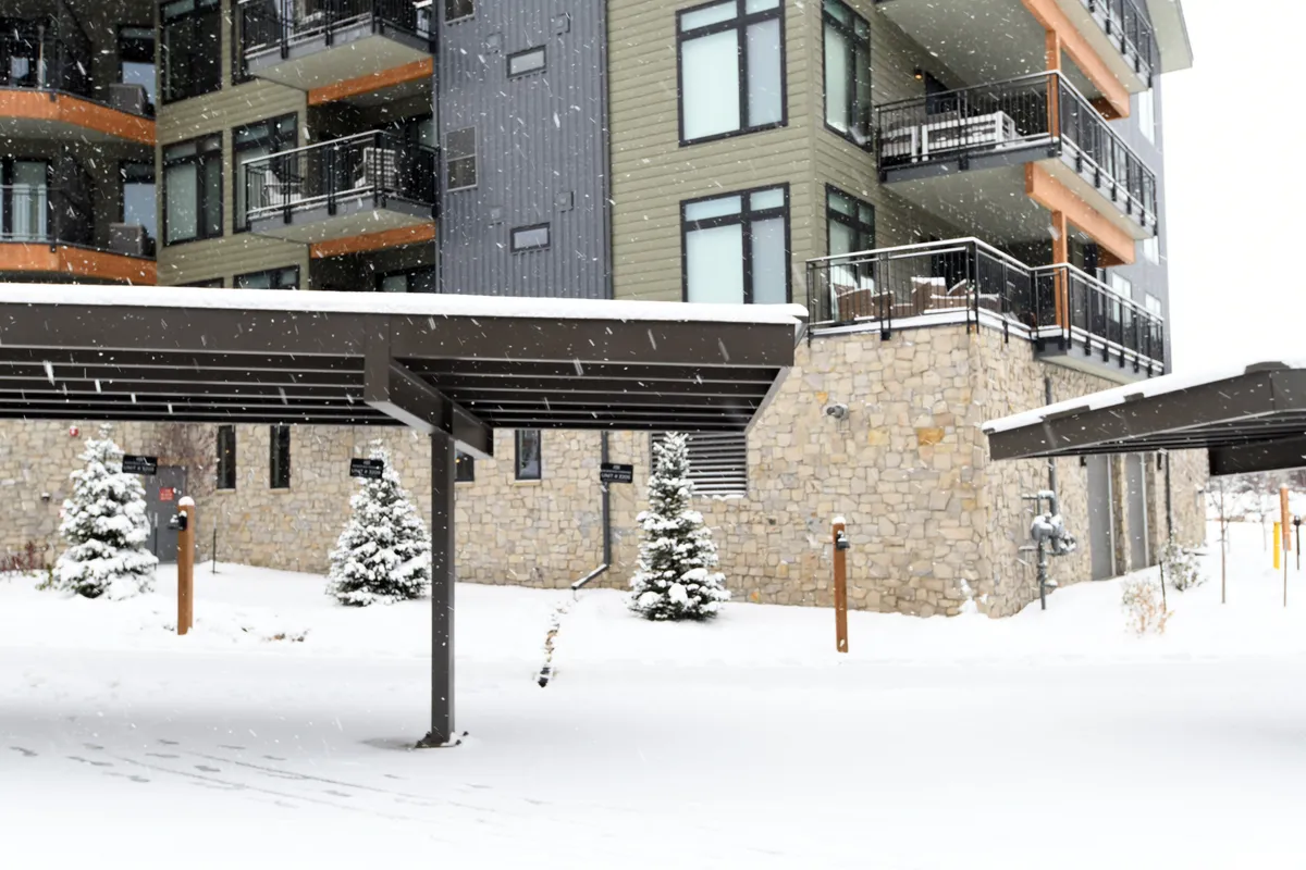 The image showcases the building's exterior blanketed in snow, with modern architecture, decorative evergreens, and carport structures in view. The tranquil, wintry landscape suggests immediate access to outdoor activities and captures the resort atmosphere perfectly.