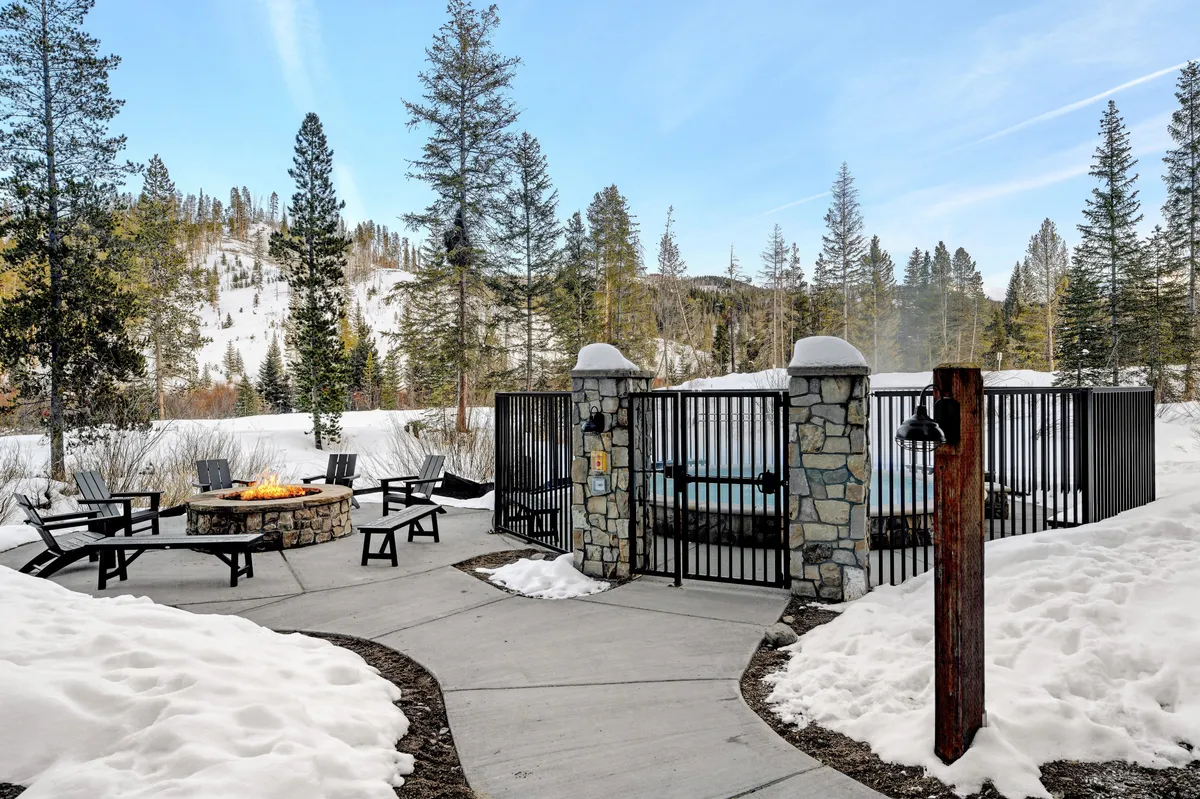 An inviting exterior community space featuring a glowing fire pit and a steaming hot tub, surrounded by snow and trees. The area is designed for relaxation, gatherings, and connecting with neighbors in a tranquil winter setting.