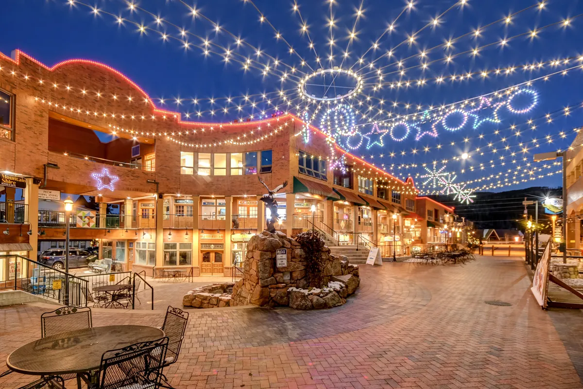A vibrant exterior scene featuring twinkling string lights, decorative stars, a brick pathway, outdoor seating, and a fountain. Boutique shops frame the scene, suggesting a lively, walkable local area perfect for dining and socializing.