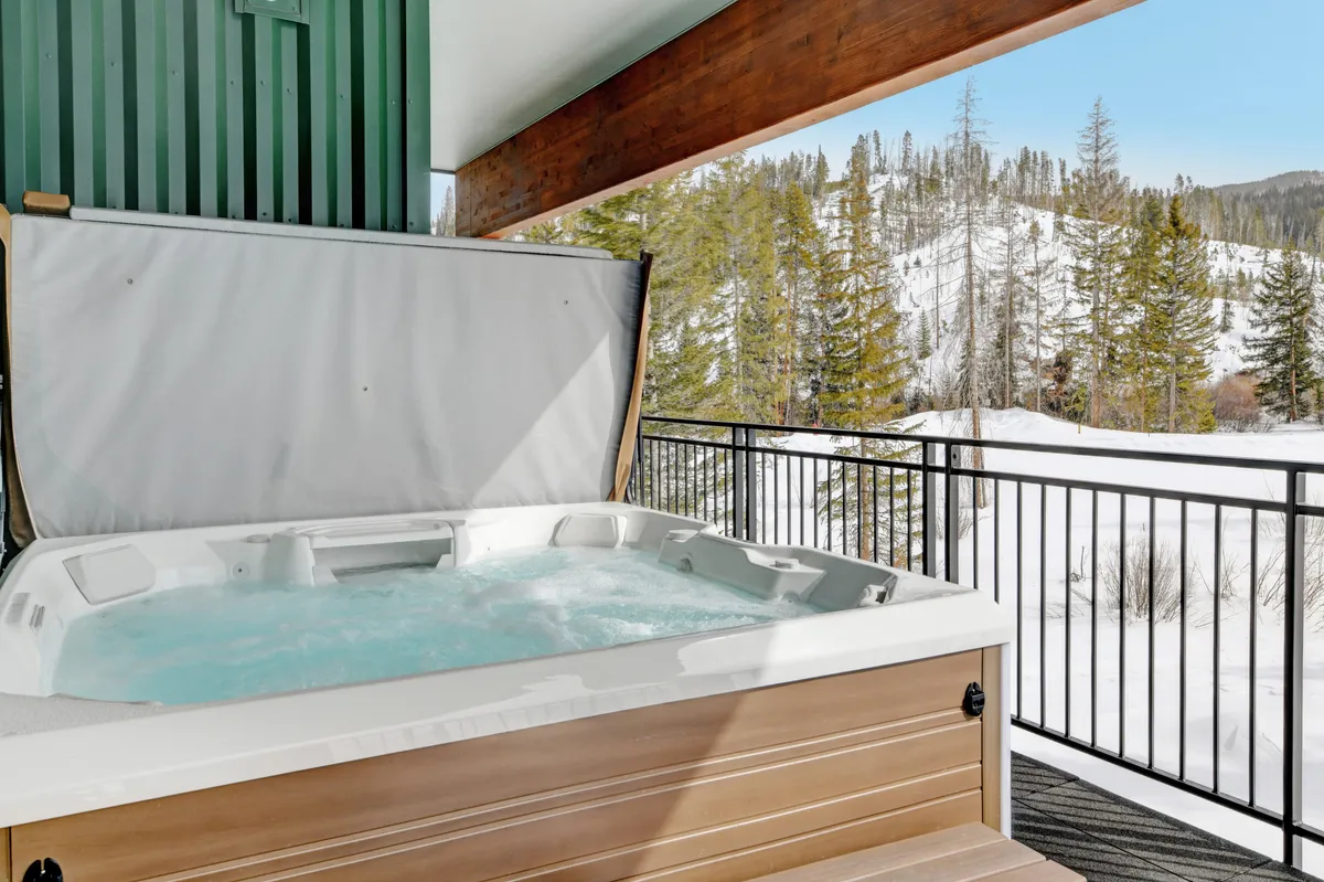 A private outdoor hot tub sits bubbling on a wooden balcony, surrounded by a snowy landscape and tall pine trees. Steam gently rises from the water, and the snow-dusted scene creates an inviting, tranquil winter atmosphere perfect for relaxing after a day on the slopes.