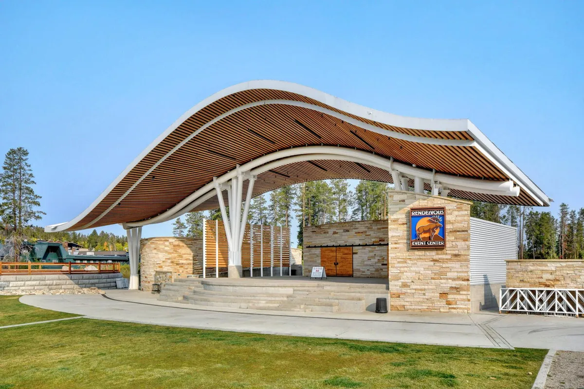 An outdoor amphitheater with a distinct curved wooden roof, stone façade, and visible event signage. The open-air setting is designed for live performances, concerts, and community events, with a welcoming layout that encourages gatherings and celebrations.